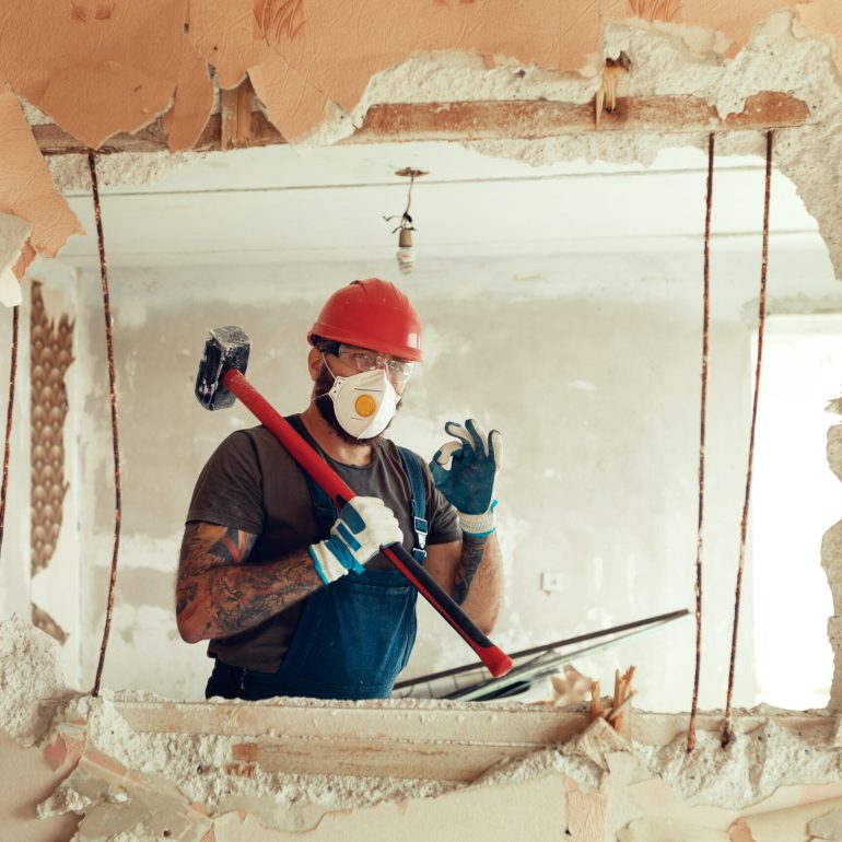 builder with a hammer in his hands breaks the cement wall The builder is dressed in a protective suit and helmet